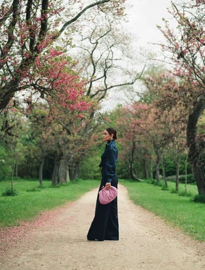 A woman in a blue jumpsuit poses in a park on a dirt path. She holds a pink clutch, with pink blossoms on the trees surrounding her.