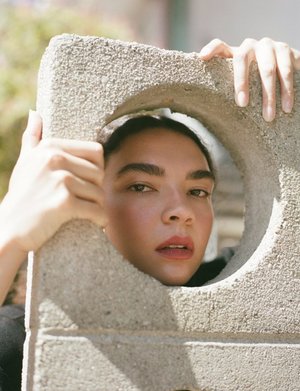 Close-up of a woman with her face framed by a concrete block with a circular hole, shot in natural light, creating a unique portrait.