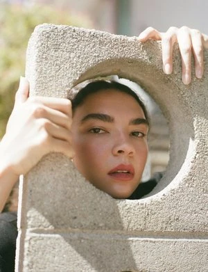 Close-up of a woman with her face framed by a concrete block with a circular hole, shot in natural light, creating a unique portrait.