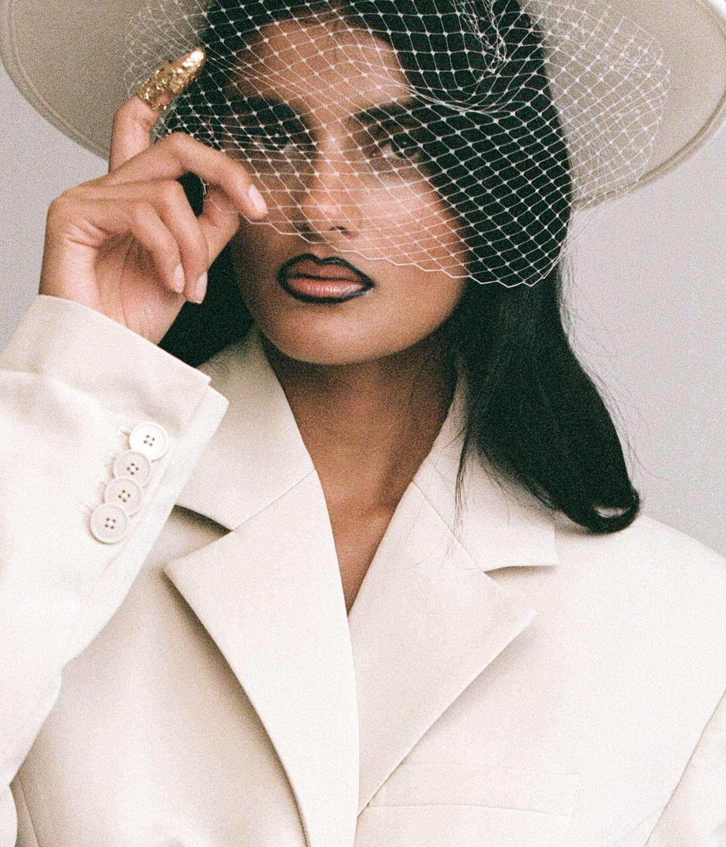 Woman in a white jacket and hat with netting poses in a close-up shot with neutral background. She wears a gold ring and dark lipstick.