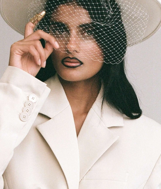 Woman in a white jacket and hat with netting poses in a close-up shot with neutral background. She wears a gold ring and dark lipstick.
