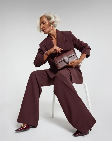 An elderly woman with silver hair wearing a matching maroon pantsuit and holding a dark red handbag while seated on a white chair against a light background.