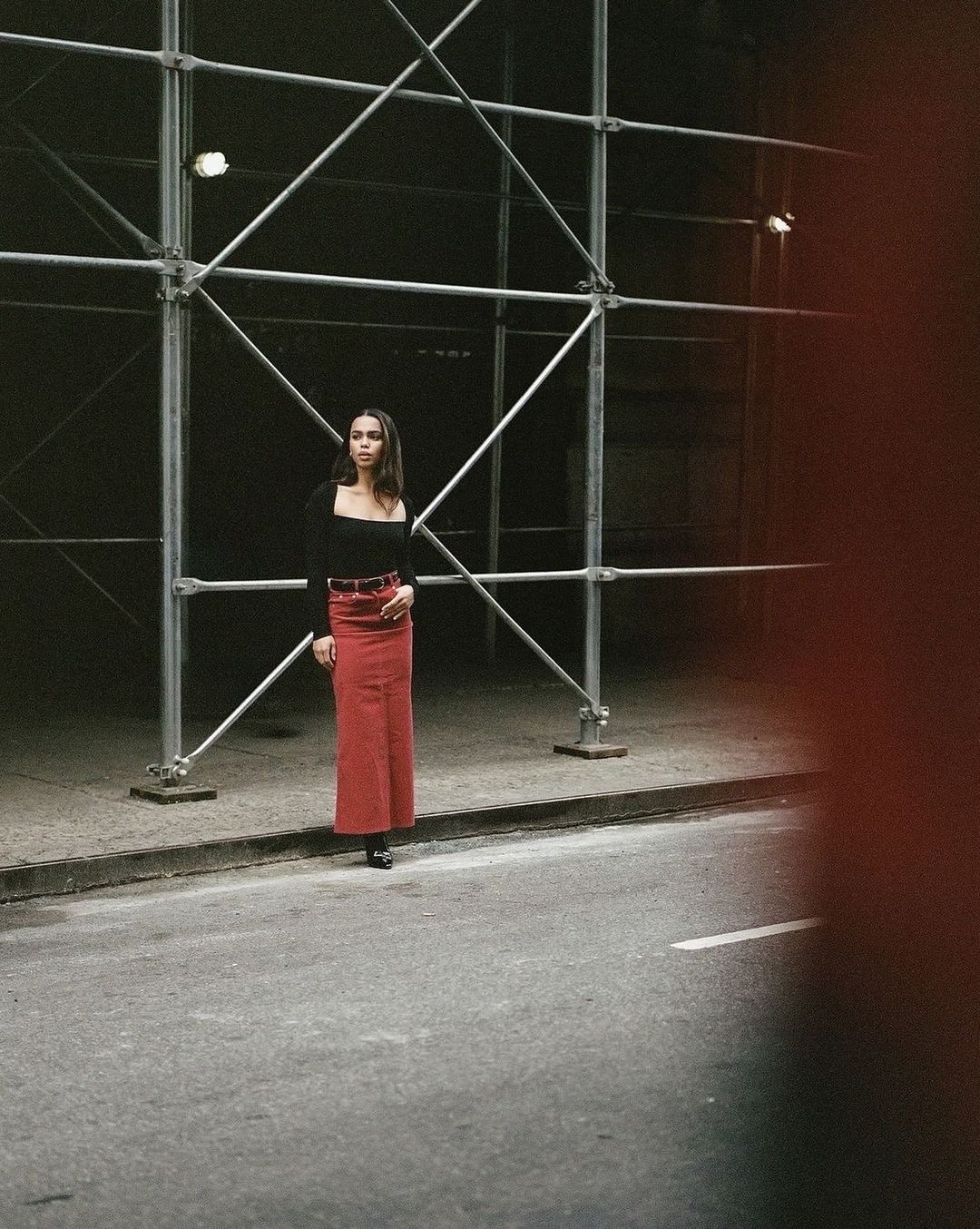Woman in red skirt and black top stands by scaffolding on a city street at night, illuminated by street lights.