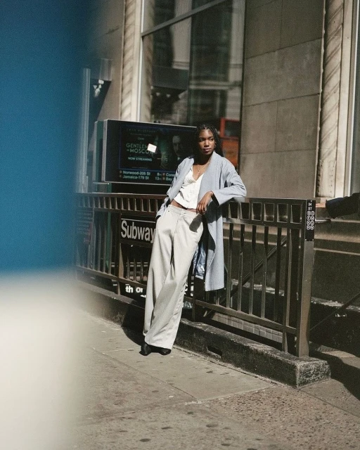 Fashionable woman in stylish coat and trousers poses near subway entrance in urban setting with sunlight. Street style and fashion editorial.