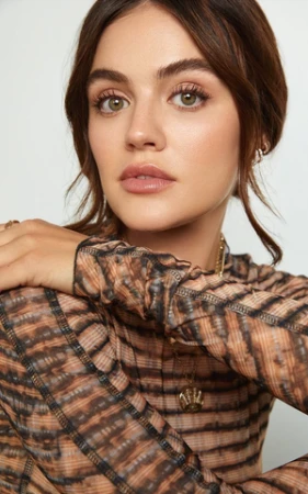 Close-up portrait of a woman wearing a patterned sheer mesh top and subtle gold jewelry against a white background.