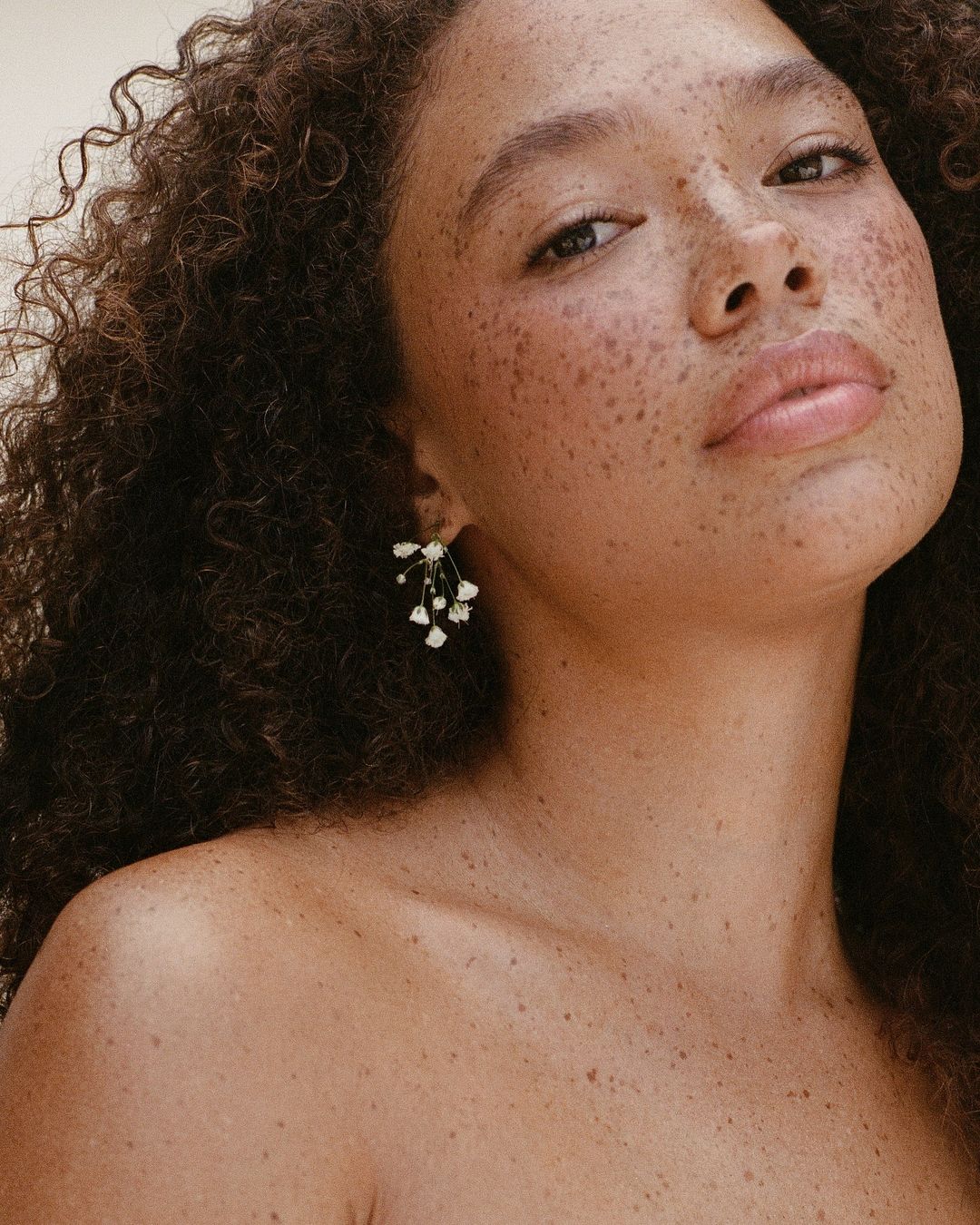Close-up shot of a woman with freckles and curly hair wearing delicate floral earrings, posing against a neutral background in soft lighting.