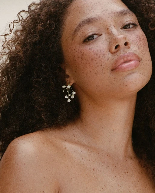 Close-up shot of a woman with freckles and curly hair wearing delicate floral earrings, posing against a neutral background in soft lighting.