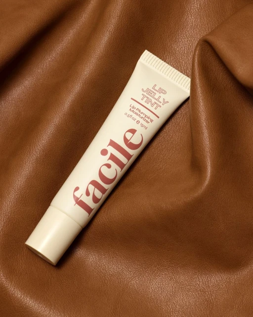 Facile Lip Jelly Tint tube on a textured tan surface in a beauty product shot. Cream-colored tube with brown text, skincare product close-up.