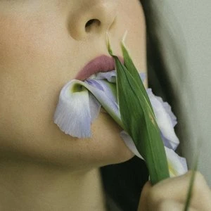 Close-up of a woman's face with pink lips holding a light blue iris flower in her mouth against a neutral background.