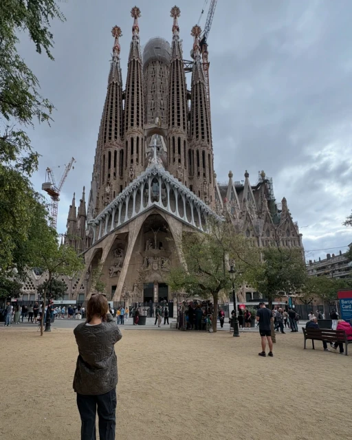 Sagrada Familia basilica in Barcelona, Spain, towers behind a tourist; trees and a cloudy sky in the background on a sunny day.