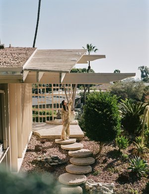 Woman in a shimmering jumpsuit poses on stone steps leading to a mid-century modern house with desert landscaping in sunlight.