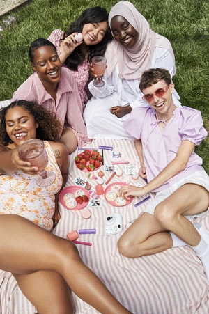 A group of diverse young adults are sitting on a striped blanket in a grassy outdoor setting, enjoying a picnic with drinks and treats.