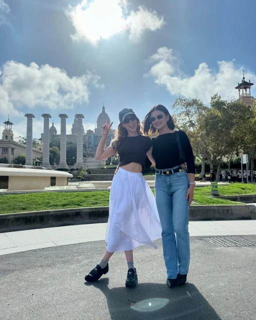 Two adult women pose on a city street in front of historic architecture under a bright cloudy sky during daytime. Barcelona.