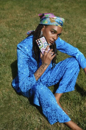 Person in vibrant blue patterned suit and silk headscarf sitting on green grass holding a decorated smartphone