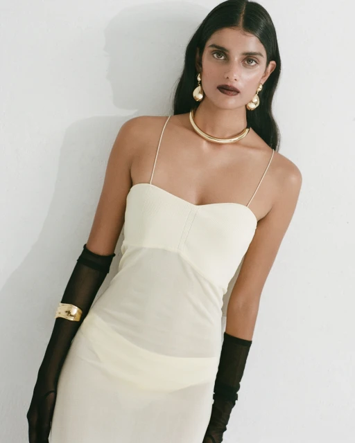 Adult woman poses indoors, wearing a cream-colored dress, sheer black gloves, and gold jewelry including earrings and a choker.
