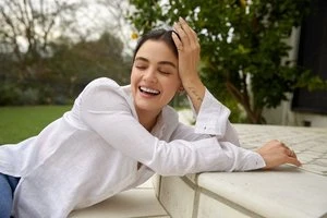 A smiling adult female with dark hair and a light complexion leans against a wall outdoors, wearing a white linen shirt and jeans.