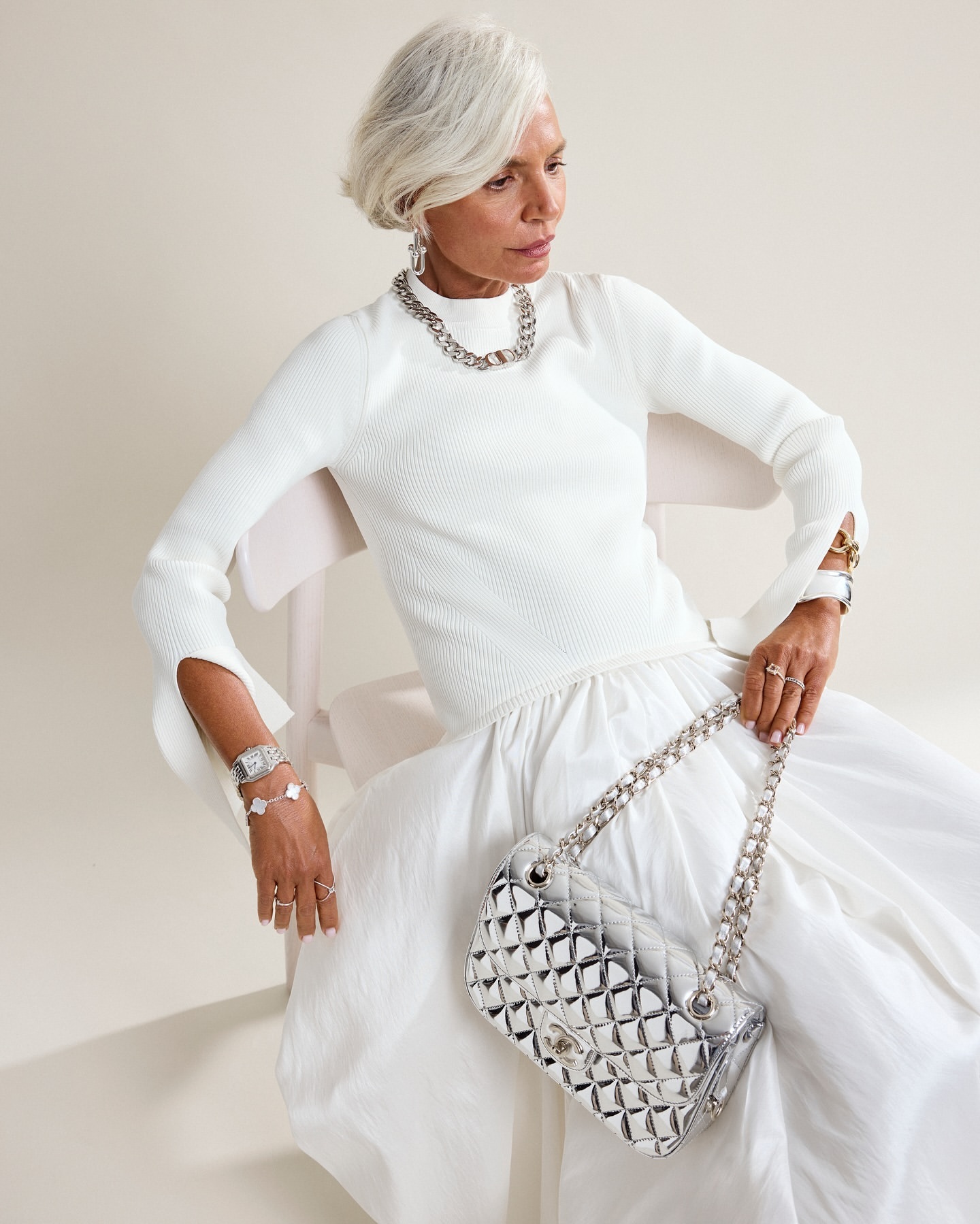 Mature woman in white outfit with silver chain bag poses in studio. Elegant jewelry and silver accessories complete her sophisticated look.