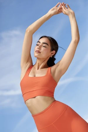 Woman in orange athletic top with arms raised against a clear blue sky