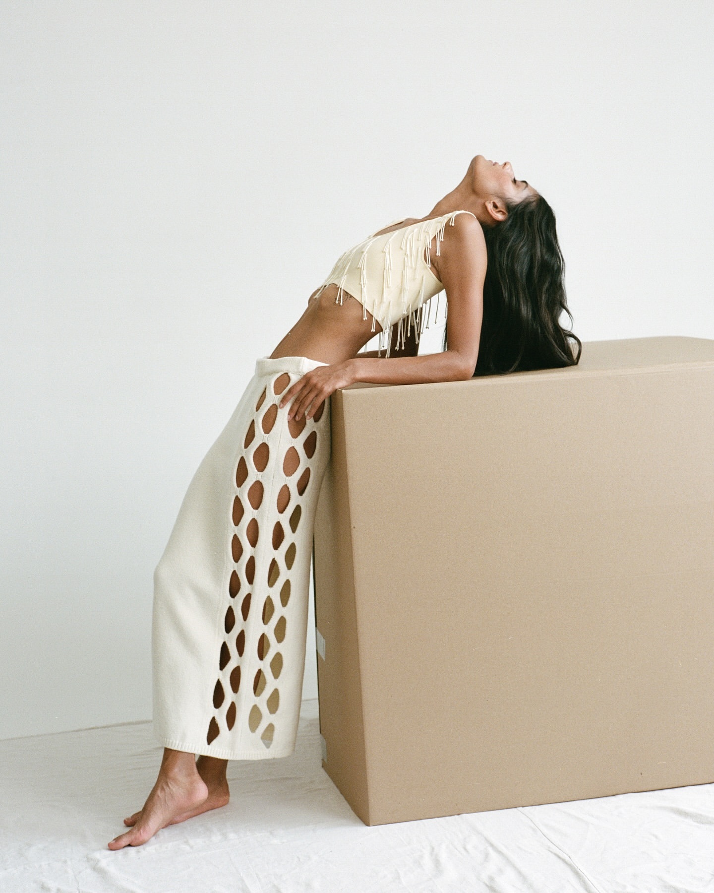 Woman in a cream-colored fringed crop top and cut-out skirt, leaning on a large tan box in a light studio setting.