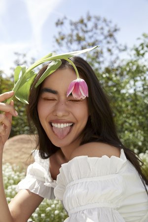 Smiling woman with a pink tulip over her eye in an outdoor setting with a blue sky and lush greenery, creating a vibrant and playful scene.
