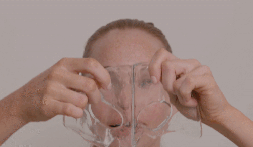 Adult female holding a clear plastic face shield near her face with a neutral expression against a plain light gray background.