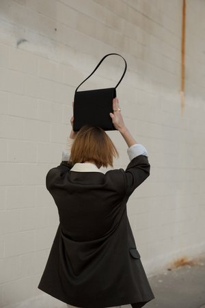 A woman in an oversized blazer holds a black handbag above her head outdoors against a light-colored brick wall, facing away from the viewer.