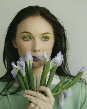 Close-up portrait of a woman with green eyeshadow holding a bunch of light blue irises against a neutral background.