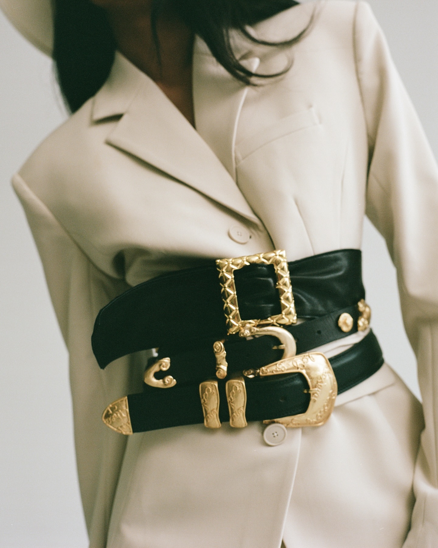 Fashionable waist-cinching black belt with ornamental gold buckles on a cream-colored blazer in a studio setting. Focus on the accessory.