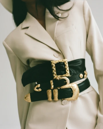 Fashionable waist-cinching black belt with ornamental gold buckles on a cream-colored blazer in a studio setting. Focus on the accessory.