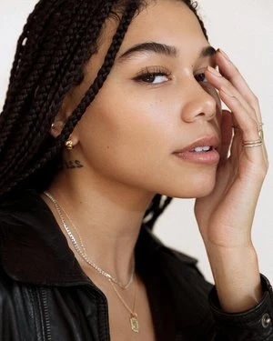 Close-up portrait of a woman with braided hair wearing layered gold necklaces and earrings against a plain white background