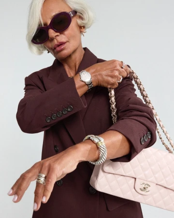 An elderly woman wearing a brown blazer, purple sunglasses, and silver jewelry holds a quilted pink handbag against a plain background.