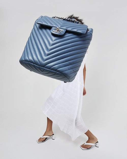 A large blue chevron quilted Chanel handbag obscures the upper body of a woman wearing a white dress and white heeled sandals.