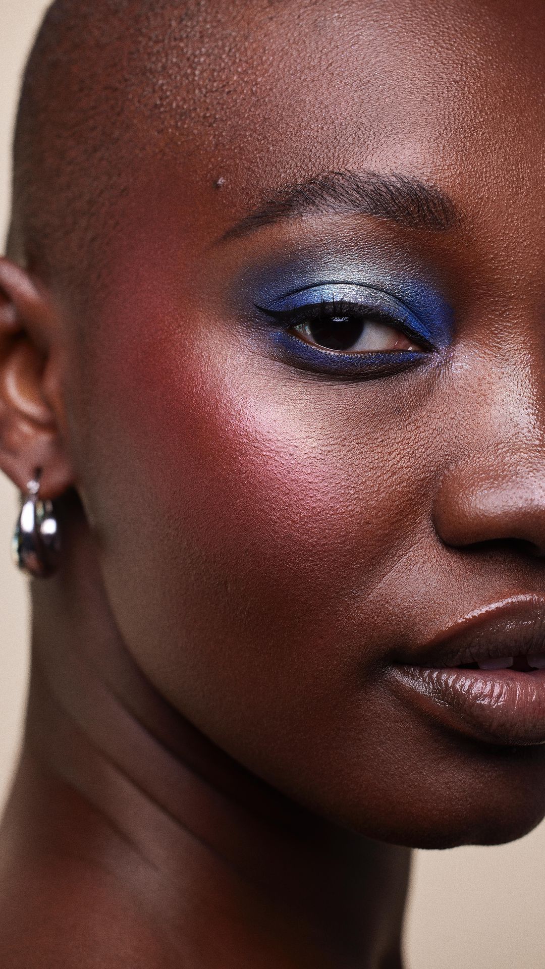 Close-up portrait of a Black woman with short hair, wearing blue eye shadow and hoop earrings, against a neutral background.