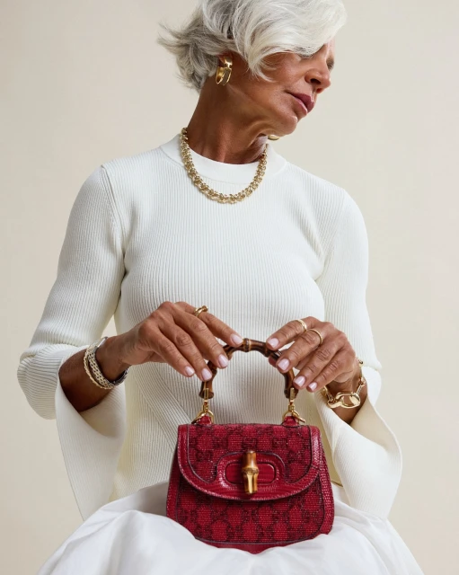 Elegant older woman presents a red Gucci handbag, complemented by gold jewelry and a white ribbed top, set against a neutral backdrop.