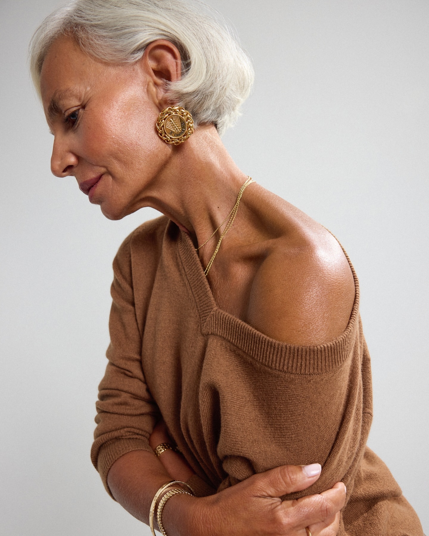 Elegant older woman with short gray hair poses in a close-up studio shot wearing a brown sweater and gold jewelry against a gray backdrop.