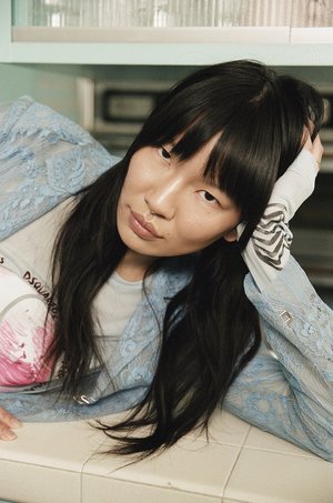 Portrait of a young adult woman with long dark hair in a light blue lace jacket and T-shirt leaning on a white tiled surface.