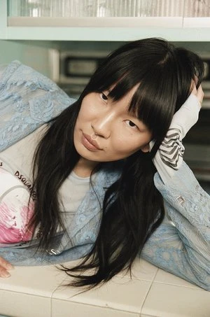 Portrait of a young adult woman with long dark hair in a light blue lace jacket and T-shirt leaning on a white tiled surface.