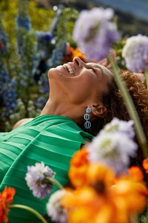 Happy woman with curly hair lays in a field of colorful flowers. She wears a green pleated top and long earrings, under natural sunlight.