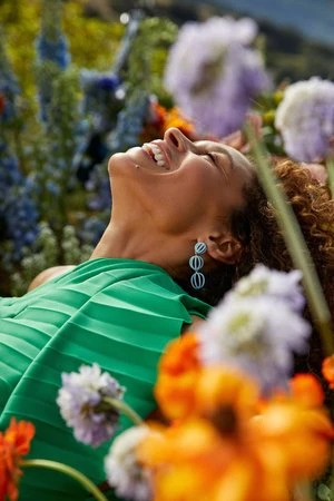 Happy woman with curly hair lays in a field of colorful flowers. She wears a green pleated top and long earrings, under natural sunlight.