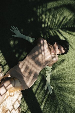 Woman in light-brown dress and light-green gloves lying on green turf with shadows of palm fronds cast across her body and the background.