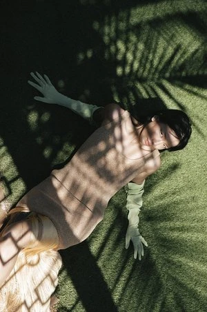 Woman in light-brown dress and light-green gloves lying on green turf with shadows of palm fronds cast across her body and the background.