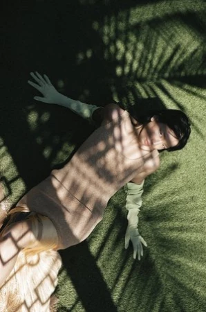 Woman in light-brown dress and light-green gloves lying on green turf with shadows of palm fronds cast across her body and the background.