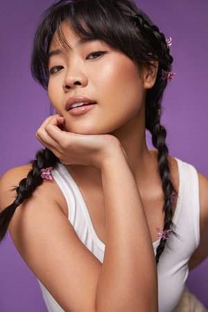 Portrait of a beautiful Asian woman with braids, her chin resting on her hand, shot in front of a solid purple studio background.