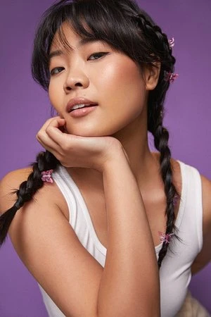 Portrait of a beautiful Asian woman with braids, her chin resting on her hand, shot in front of a solid purple studio background.