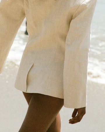 Close-up of a person wearing a beige blazer jacket outdoors near a sandy beach with ocean waves.