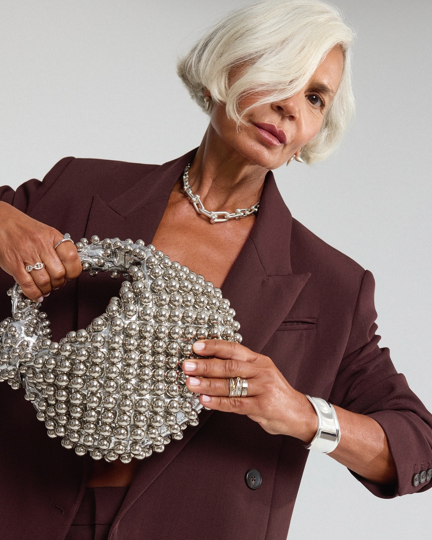 Older woman in a plum blazer holding a beaded purse, wearing rings and a chunky necklace, posing in front of a white backdrop.