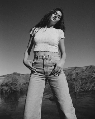 Black and white portrait of a woman wearing a white t-shirt and high-waisted denim jeans standing outdoors near water.