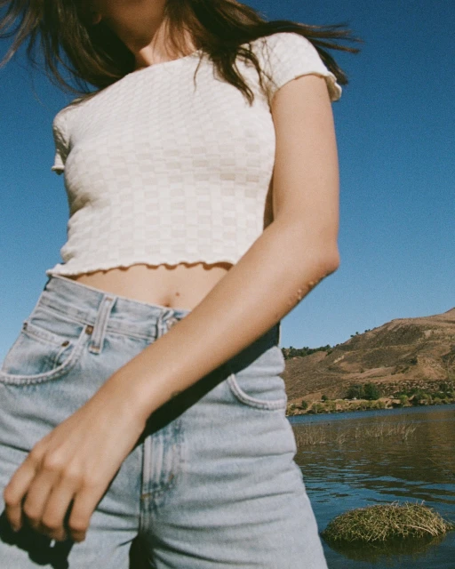 Young adult woman in jeans and a textured shirt stands outdoors near water, with a grassy mound and distant mountain backdrop.