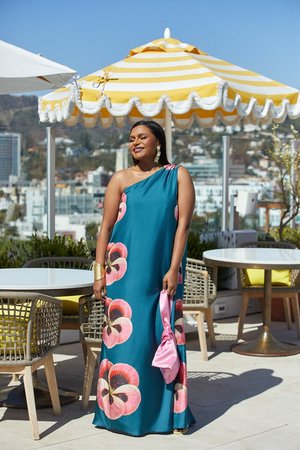 Woman in a teal one-shoulder dress with floral pattern, gold cuff bracelet, and pink purse stands on a patio with yellow umbrella.