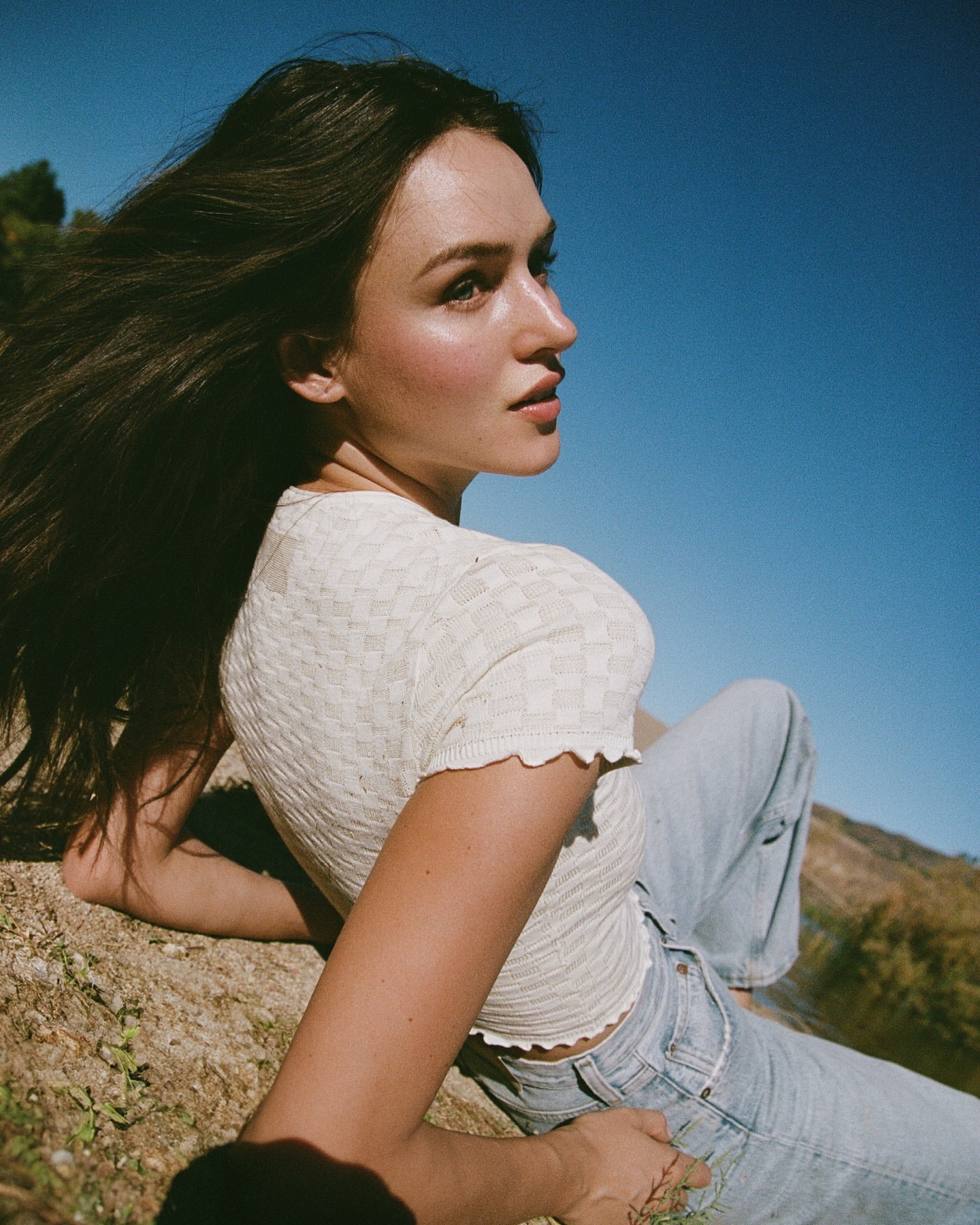 A young woman with long dark hair is lying down in a sunny field wearing jeans and a cream-colored knit shirt against a clear blue sky.